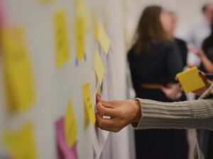A hand placing a yellow sticky note onto a wall of Post-It’s during a workshop