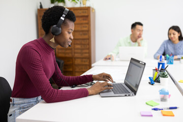 African woman focuses on collaborative writing in an office, working from a laptop and wearing headphones.